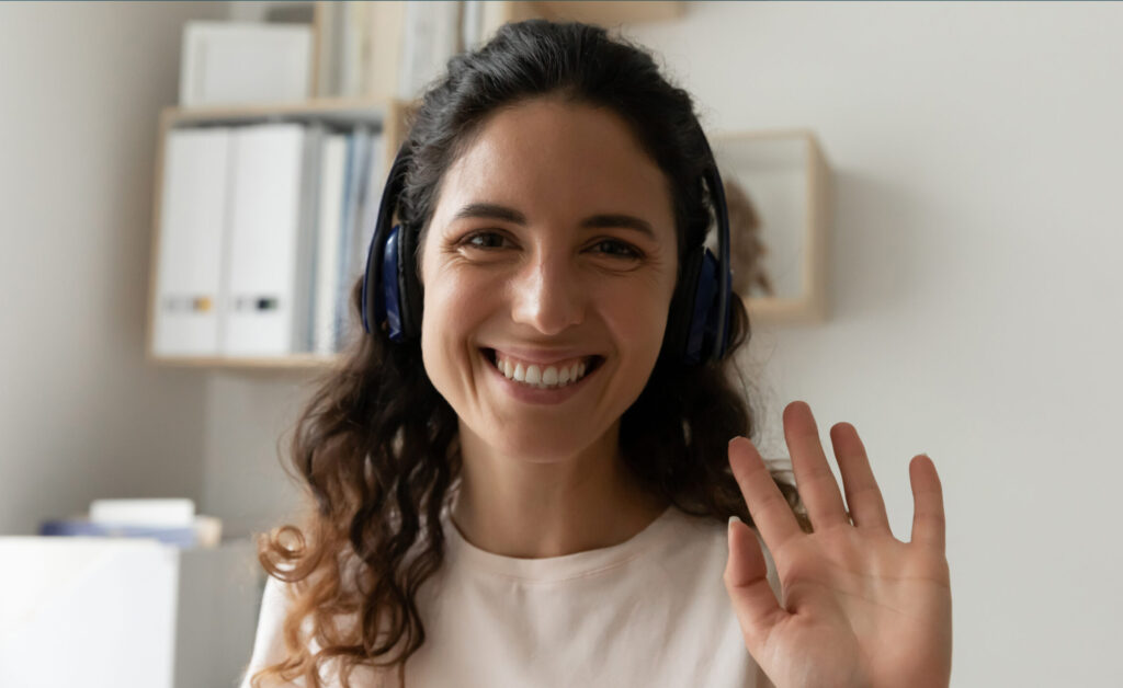 Close up photo of a young woman with headphones on looking at camera and waving hello.