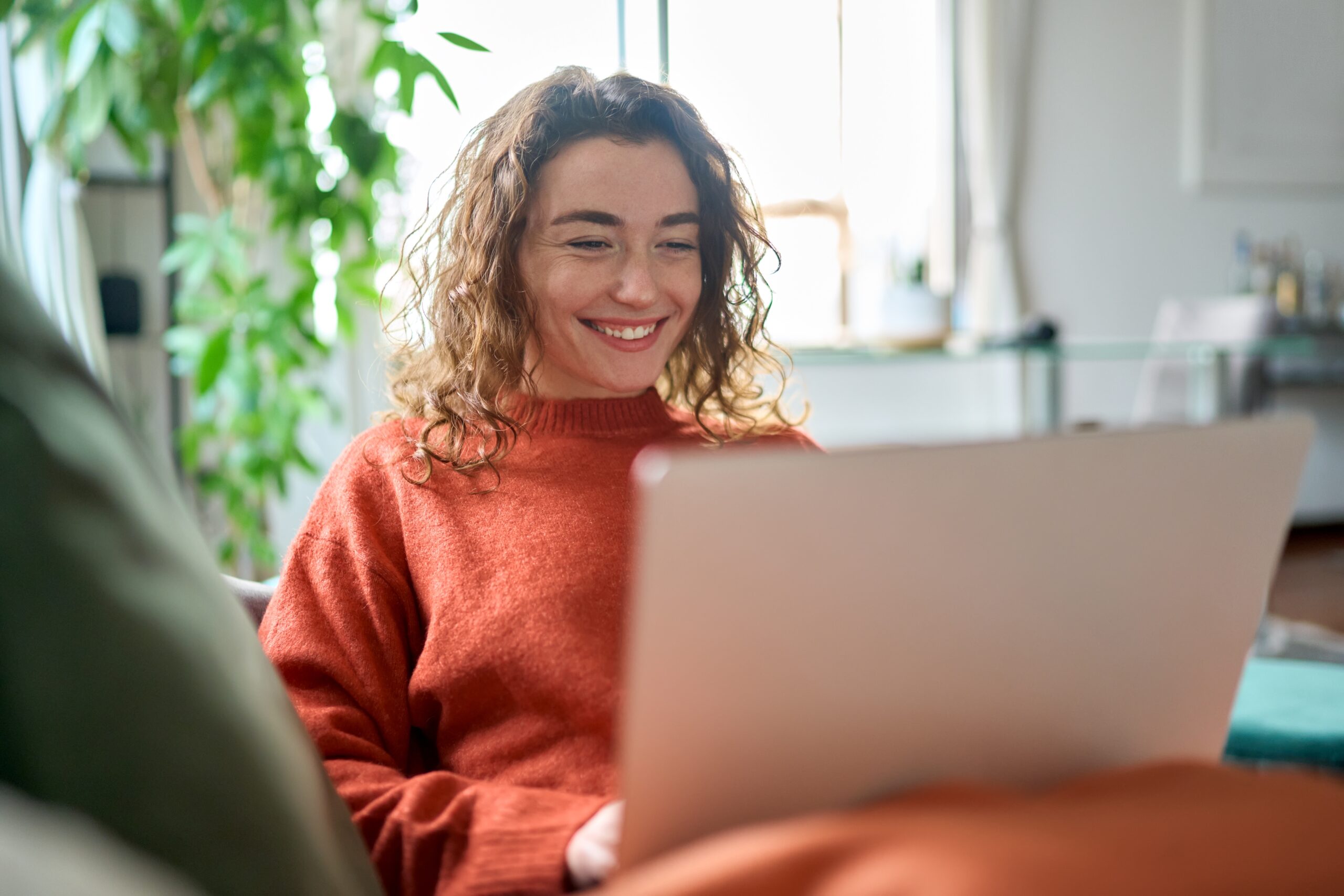 Young woman sitting indoors looking at her laptop and smiling.