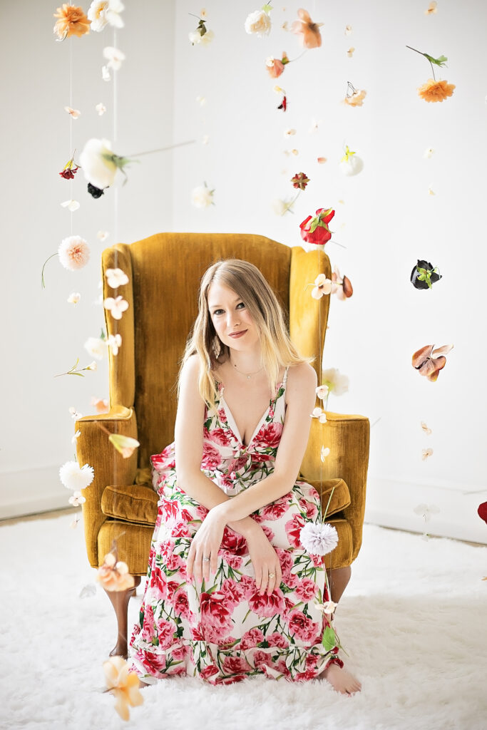 Amie, a blonde-haired white woman, sits on a yellow armchair in a colorful sundress. Paperflowers are hanging from the ceiling around her face/