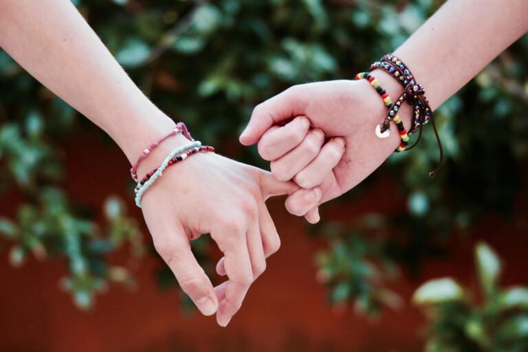 two white hands holding pinkies with bracelets on in front of a background of greenery