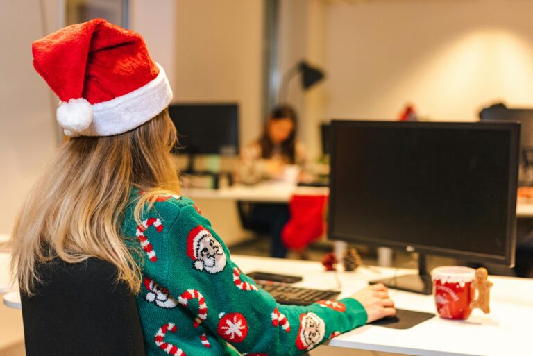 woman in an office at a desk with a computer wearing a holiday sweater and santa hat