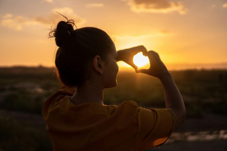 backlit woman making a heart shape with her hands in front of a sunset/sunrise