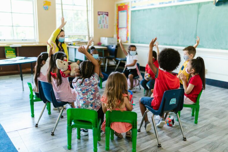 a teacher with a mask on, children in a circle around her, some of them have their hands raised