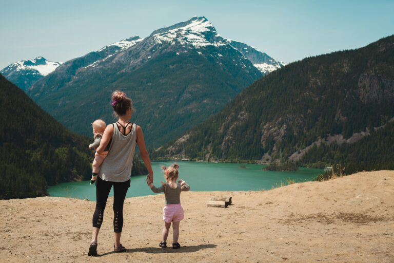 young woman with baby on her hip holding a toddler's hand in front of a lake and mountain